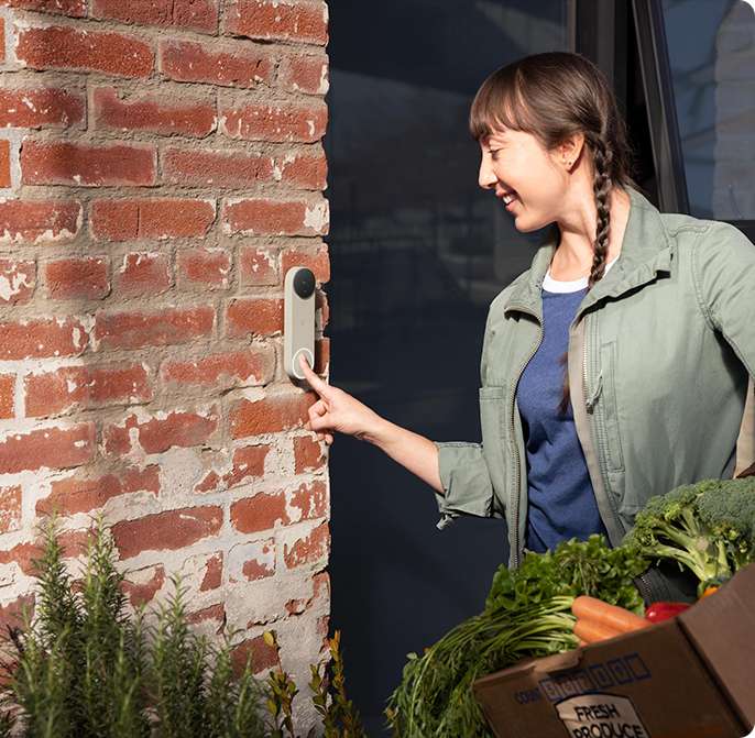 A woman pressing a Nest Doorbell on a brick wall while carrying groceries.
